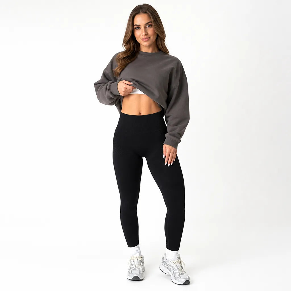 Model lifting the hem of a gray cropped sweatshirt to show the high waistband of black seamless leggings, front view. 5