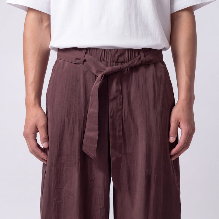 Close-up of the waist of plum-brown pants showing the elastic waistband, knotted fabric tie, front fly, and creased fabric texture. 9
