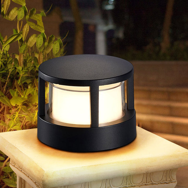 Round black post lamp with a flat cap and glowing frosted diffuser, lit on a stone pedestal beside garden plants at night. 1
