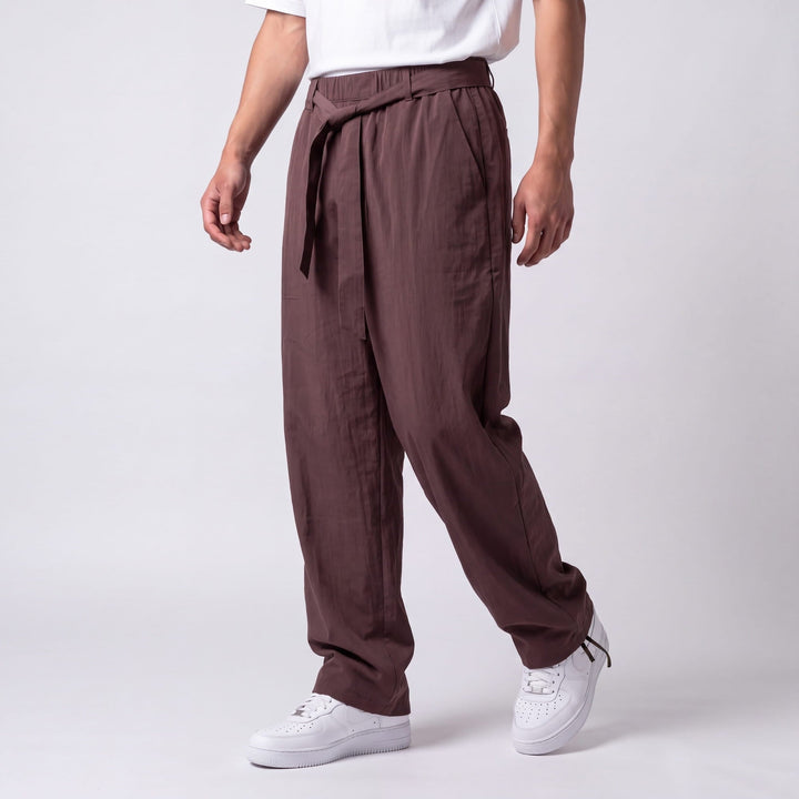 Three-quarter side view of a model mid-step in plum-brown pants with a knotted waist tie, side pocket, and ankle drawcord. 8