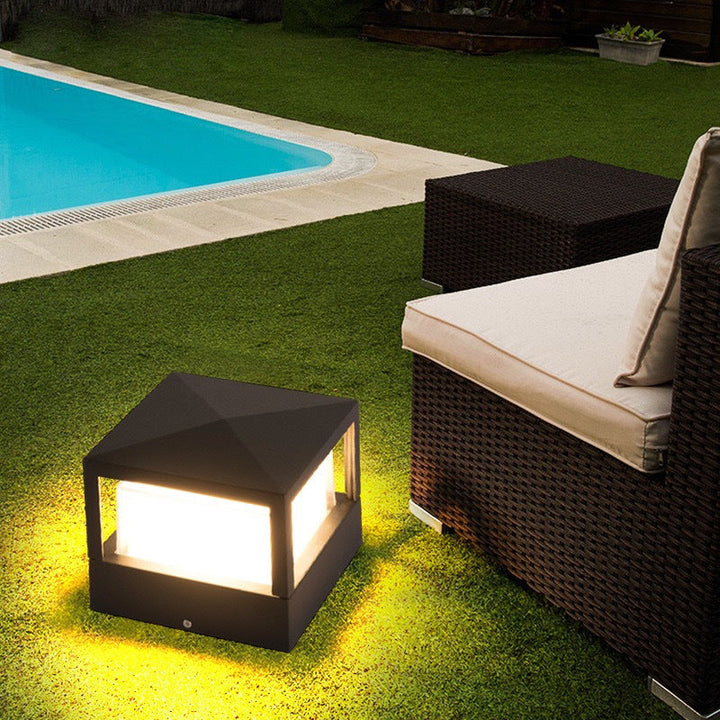 Pyramid-topped black square lamp glowing on a lawn near a swimming pool and a wicker outdoor sofa with cushions. 4