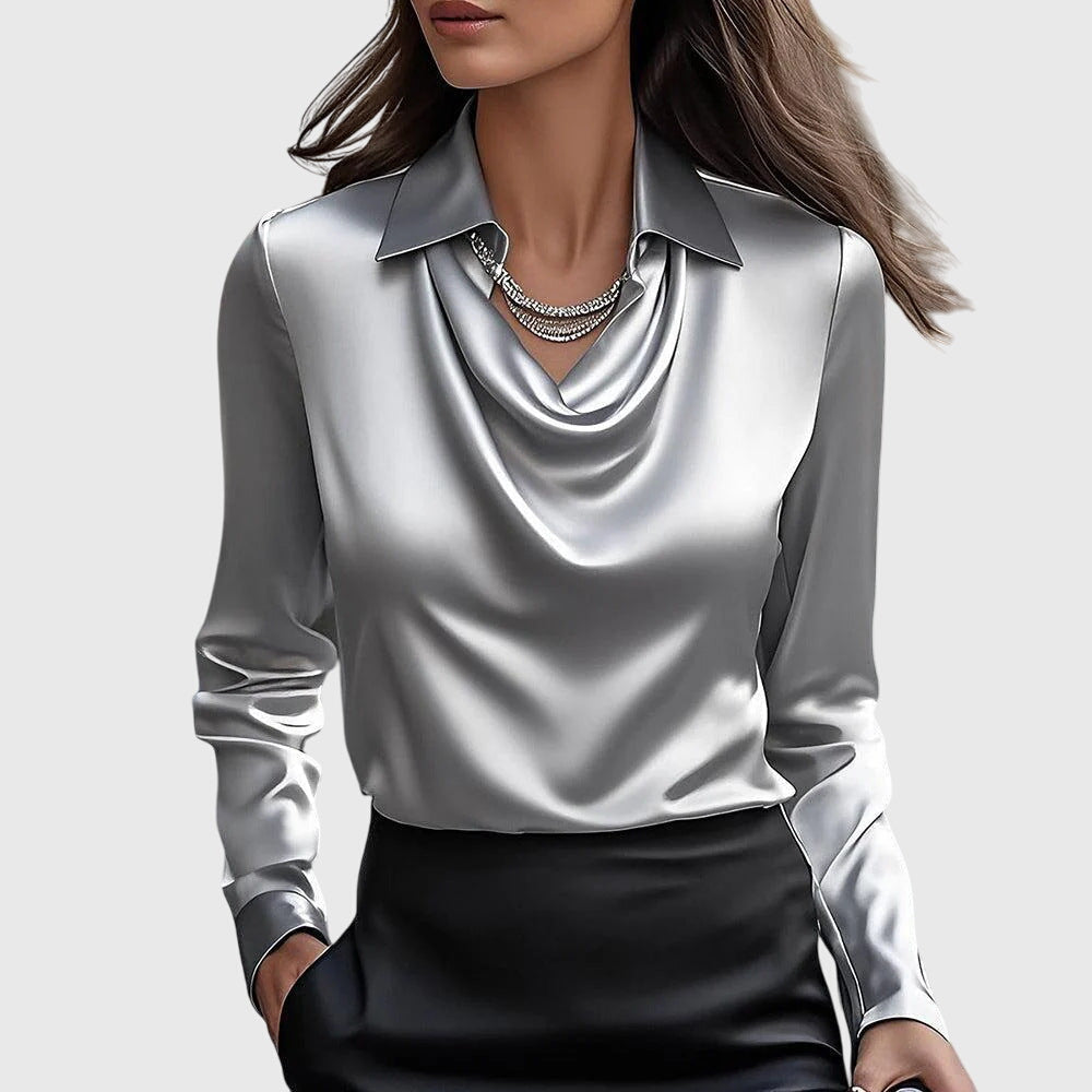 Model wearing the satin blouse in metallic silver-gray, showing detail of the cowl neck, long sleeves and a layered crystal necklace.