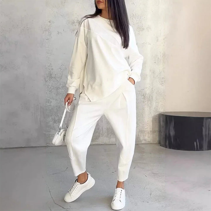 Woman wearing the white set with one hand in her pocket, holding a small white bag, standing against a concrete wall.