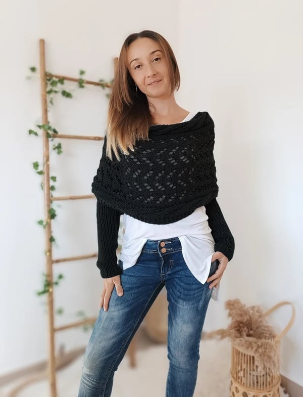 Woman poses in the black lace-knit wrap with sleeves, wearing a white top and jeans in front of a vine-wrapped ladder. 12