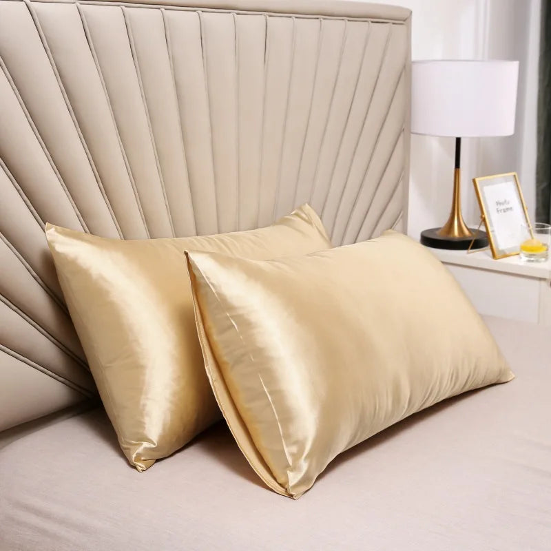 Pair of champagne gold shiny pillowcases on a bed with a pleated headboard, lamp and frame in the background. 5