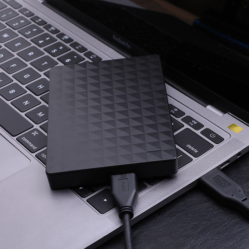 Black drive resting on a laptop keyboard, connected by a black USB cable to the laptop's side port.