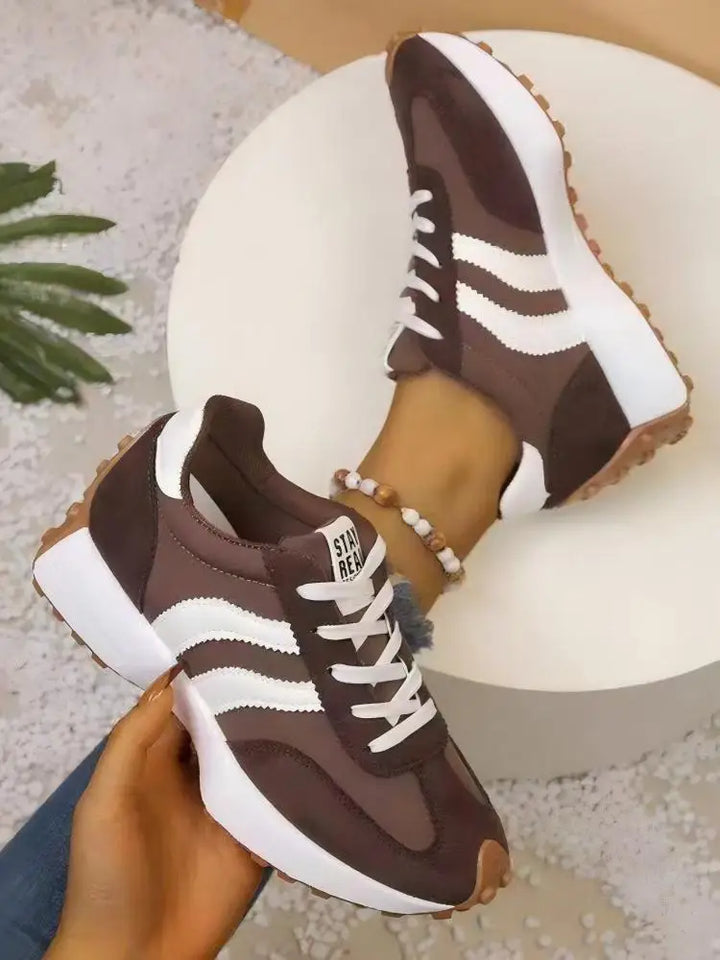 Brown trainers with white stripes held in one hand above a round white pedestal, with the other shoe worn 10