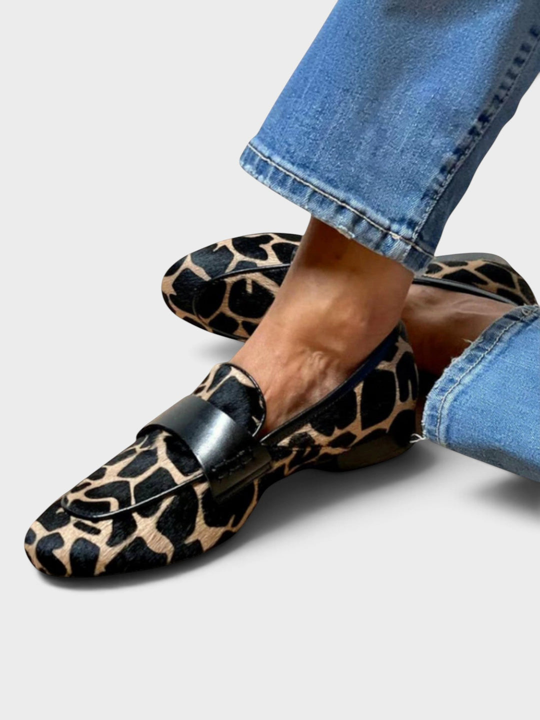 Worn leopard print loafer with black leather strap, seen from the side with ankles crossed and cuffed blue jeans. 1