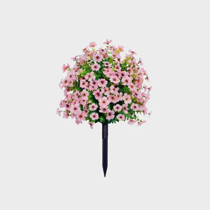 Light pink artificial flower bush with green foliage on a black ground stake against a plain background.
