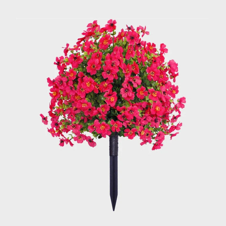 Hot pink artificial flower bush with green foliage on a black ground stake against a plain background.