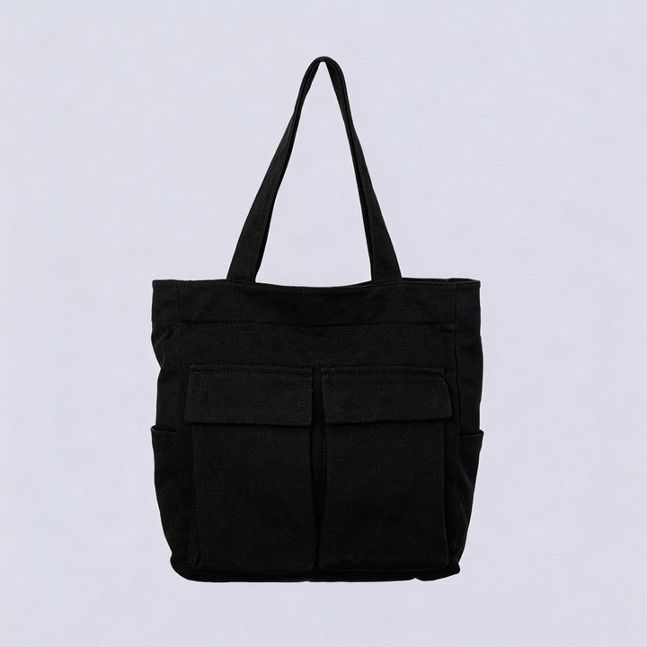 Black canvas tote bag with two front flap pockets, side pockets and a single shoulder handle, shown against a plain light background. 1