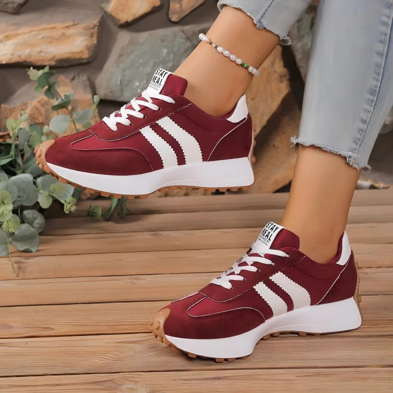 Burgundy trainers with white stripes and a lace-up front, worn on a wooden deck beside green plants 11