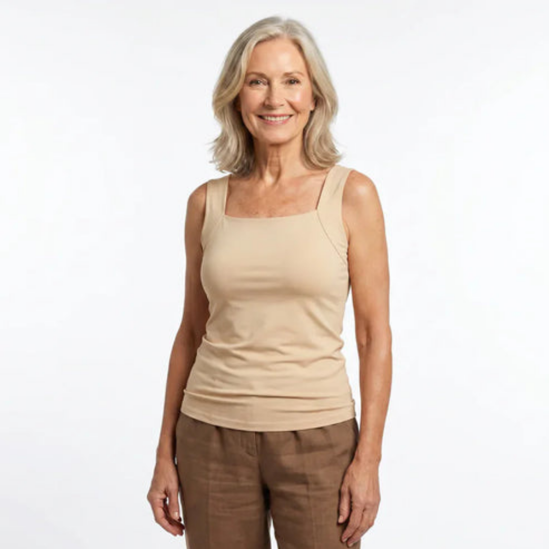 Smiling woman with shoulder-length gray hair stands facing front in a beige square-neck tank top and brown linen pants. 1
