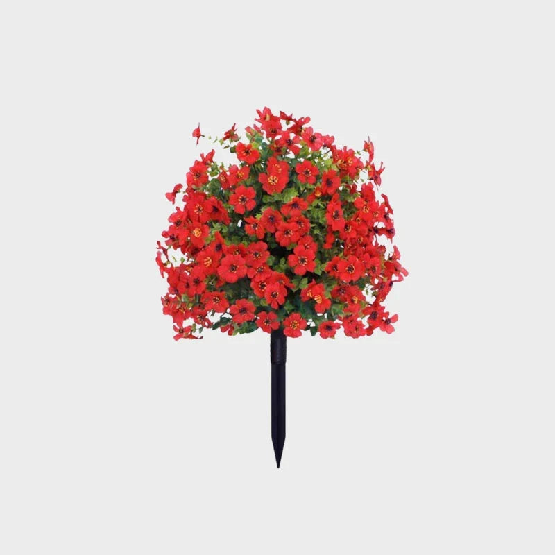 Bright red artificial flower bush with dark green foliage on a black ground stake against a plain background.