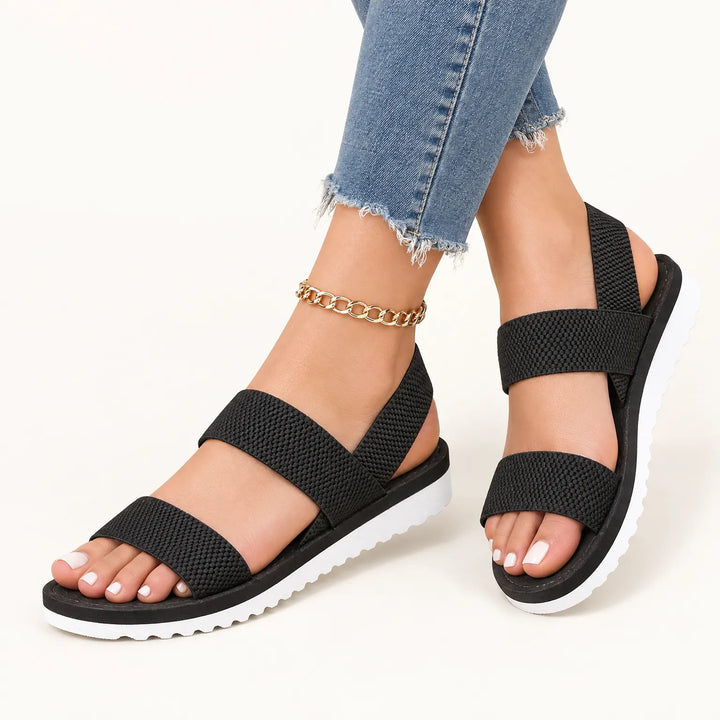 Black textured elastic double-strap sandals with white ridged soles, worn with frayed jeans and a gold chain anklet on white 11