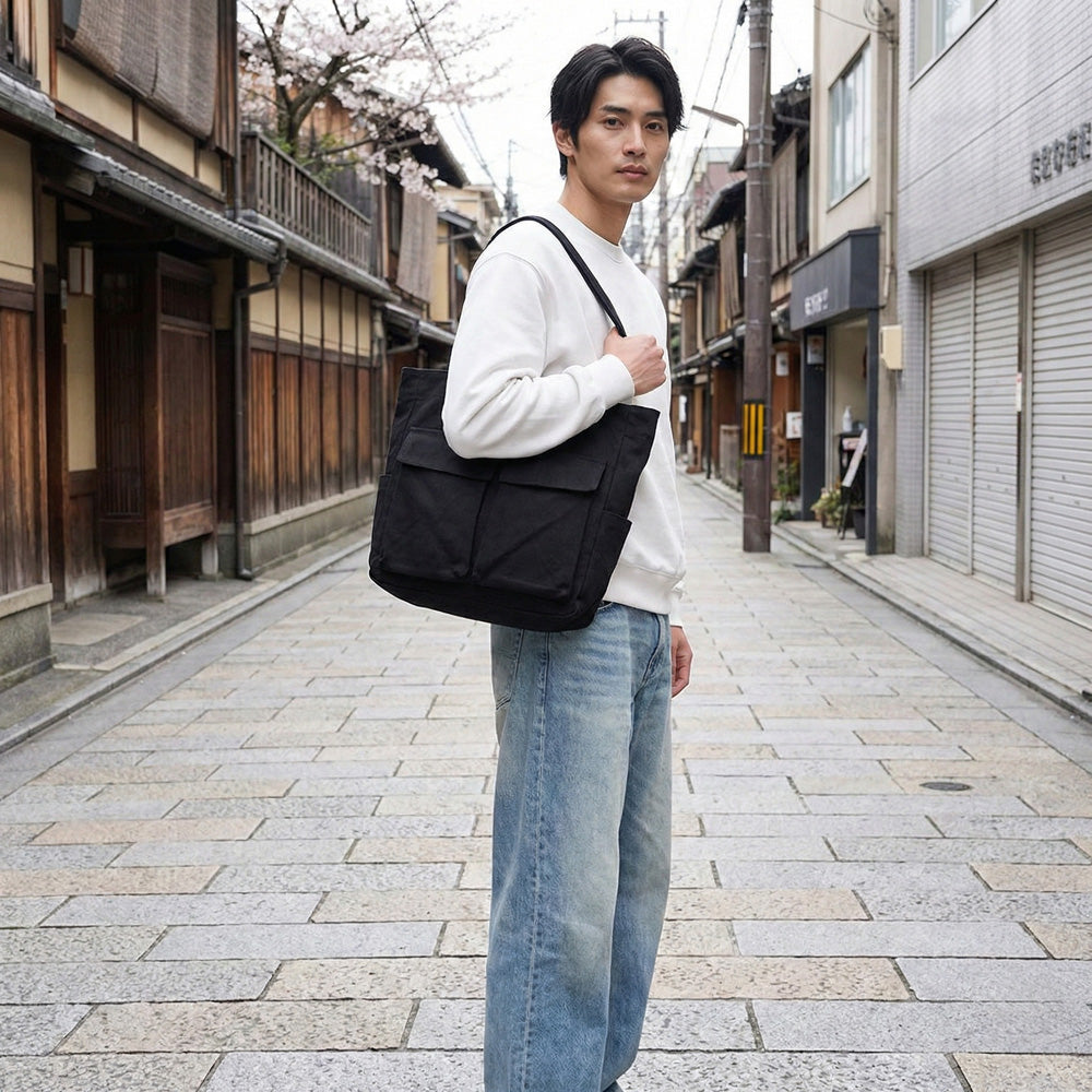 Man in a white sweatshirt and light blue jeans carries the black tote on one shoulder while standing on a stone-paved street. 2