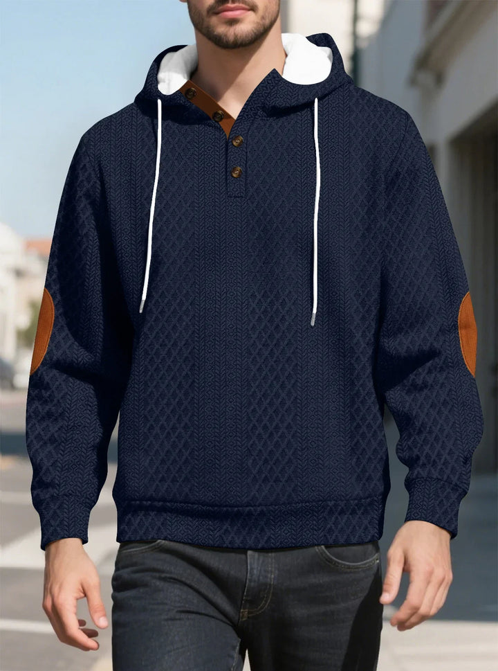 Front view of a man walking in a navy diamond-textured hoodie with brown elbow patches on a sunny street 13