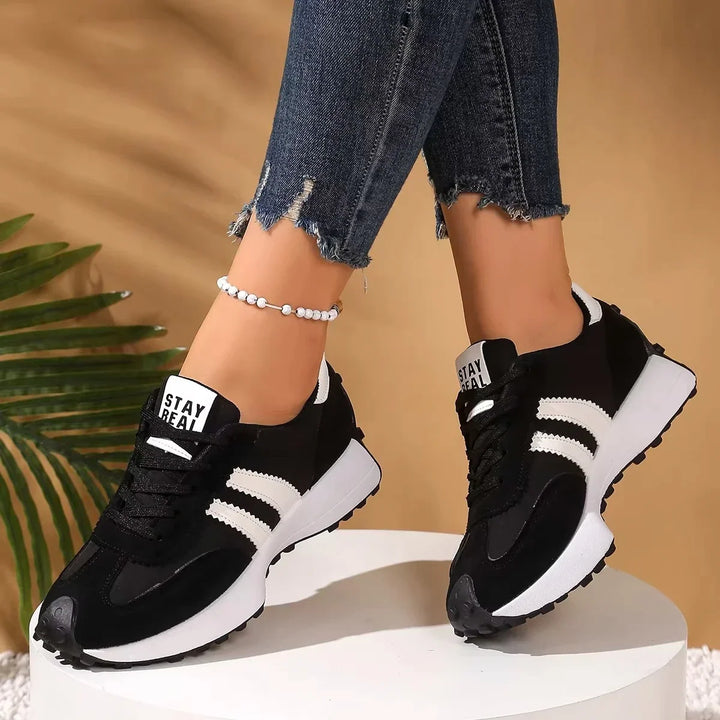 Black platform trainers with white stripes worn standing on a white pedestal, front three-quarter view with heel tab 14