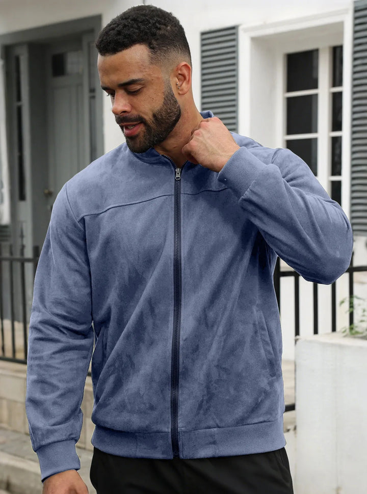 Bearded man in a blue zip-up jacket, hand raised to his collar, standing near a white house 15