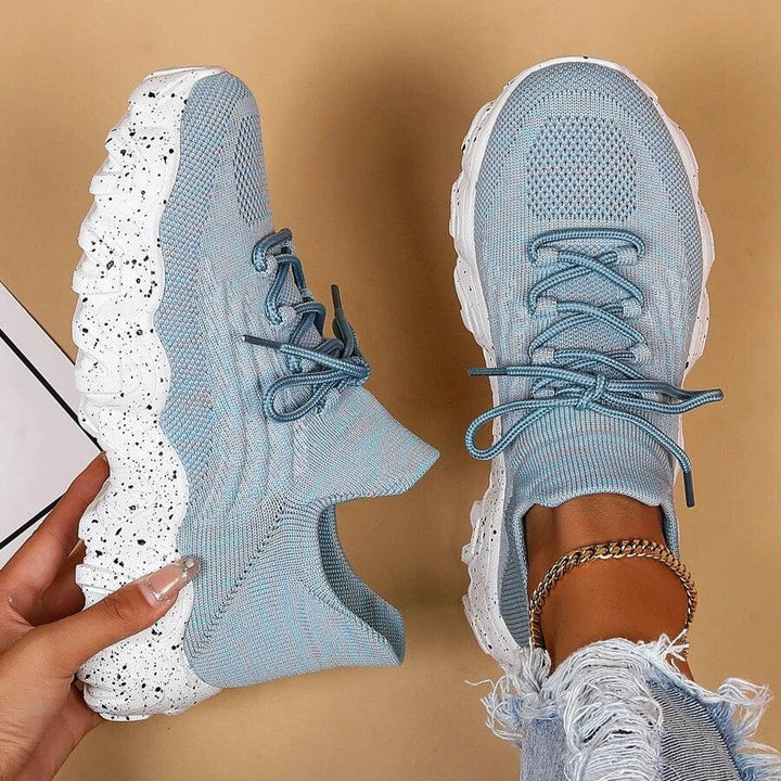 Another view of light blue knit sneakers, one held in hand showing the speckled white sole and one worn with frayed jeans.