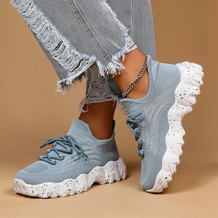 Another view of light blue knit sneakers and ripped jeans, one foot raised to show the white speckled sole from the side.