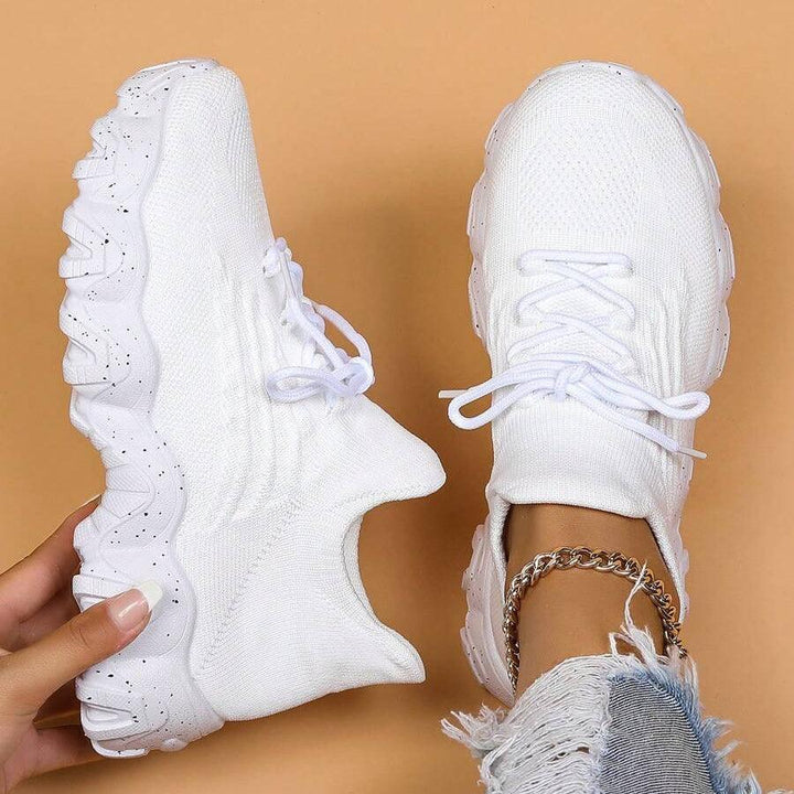 Another view of white knit sneakers, one held in a manicured hand showing the sole and one worn with ripped jeans.