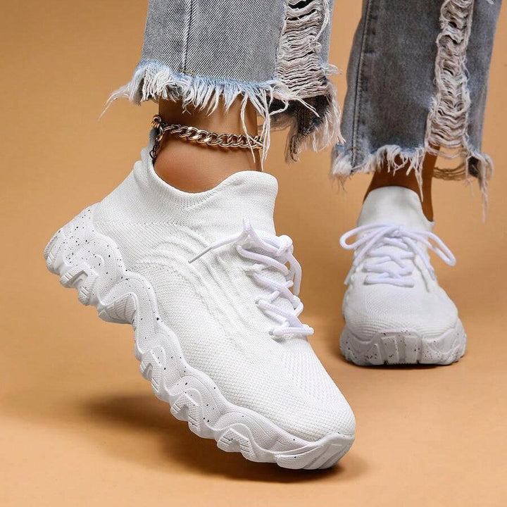 Another view of white knit sneakers with ripped denim and a gold ankle chain, shown from the front.