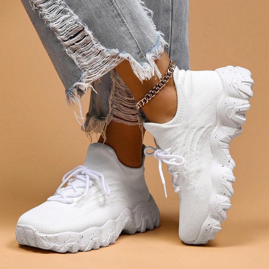 Another view of white knit sneakers with one foot raised on its side, showing the thick speckled sole and lacing.