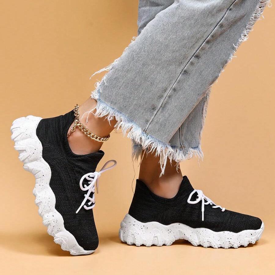 Model standing in black knit sneakers with white laces and speckled white soles, with one shoe tilted to show its profile. 34