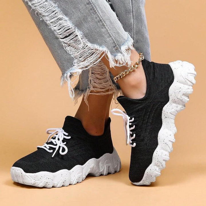 Model with crossed legs in black knit sneakers with white laces, showing the tall speckled white sole from the side. 35
