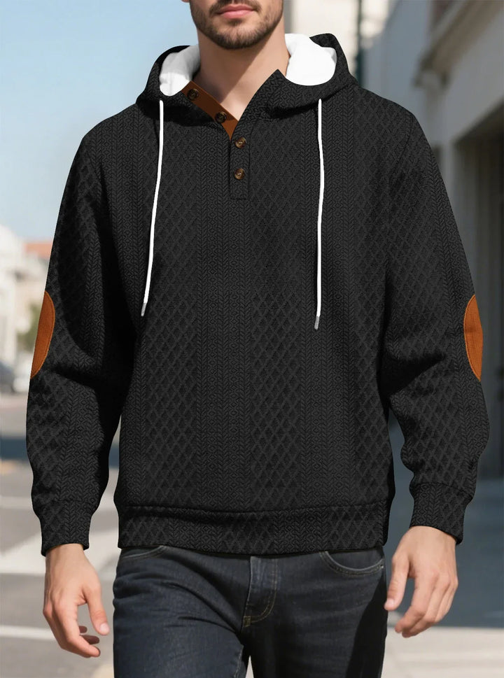 Front view of a man walking in a black diamond-textured hoodie with brown elbow patches on a sunny street 16