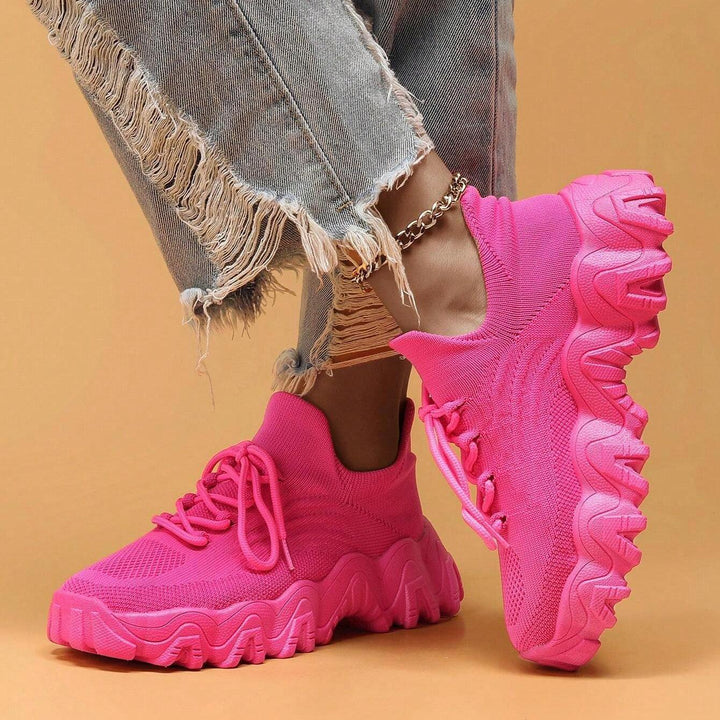 Model crossing her legs in hot pink knit sneakers, showing the tall wavy sole from the side and the gold ankle chain. 25