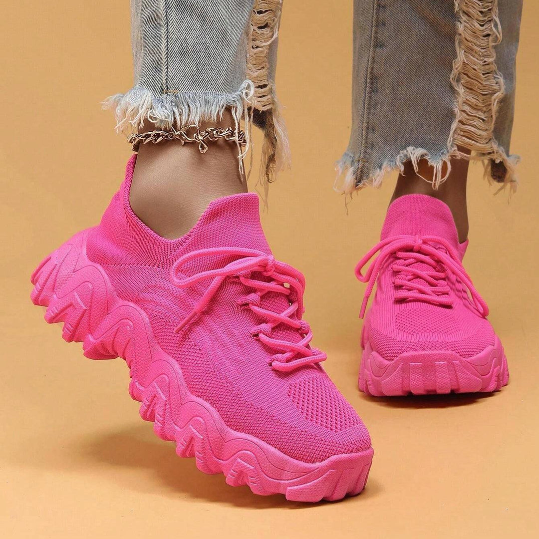 Model walking in hot pink knit sneakers, front three-quarter view showing laces, mesh toe panel and thick ridged sole. 24