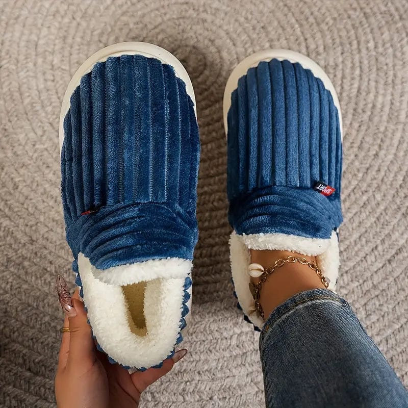 Top view of deep blue ribbed slippers on a woven rug, one held in a hand and one worn with a shell anklet.