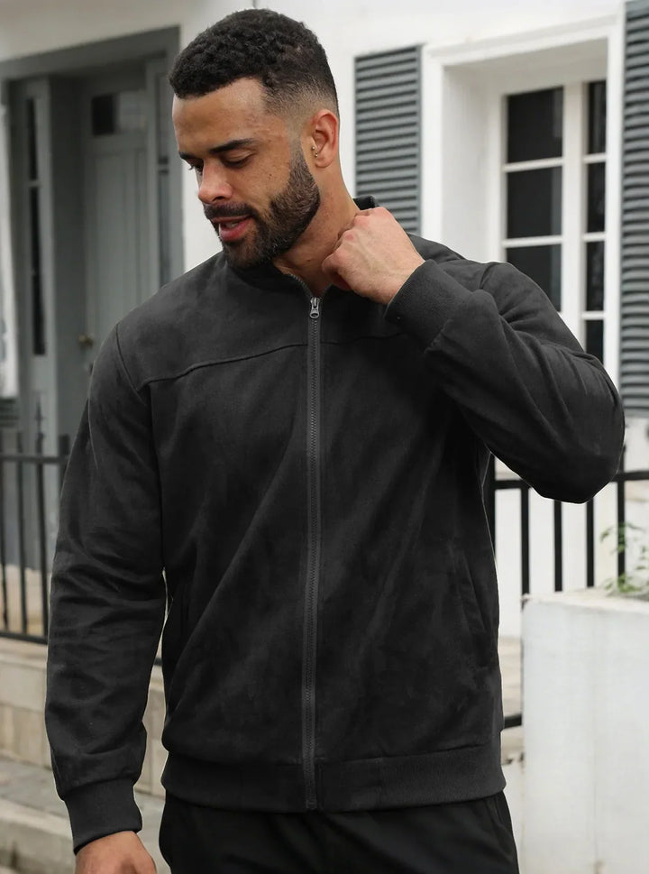 Bearded man in a black zip-up jacket with ribbed cuffs, hand at his collar, outside a white house 19