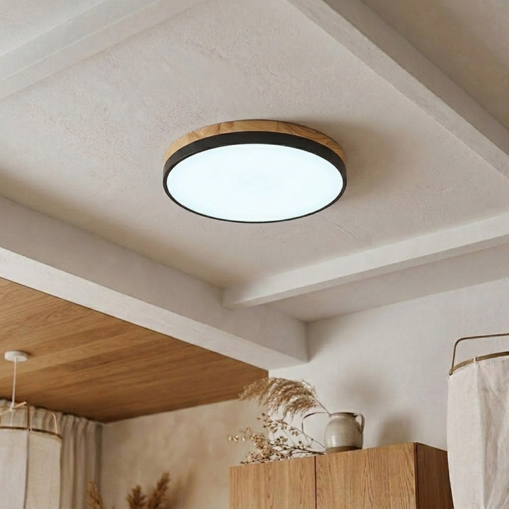 Black-banded disc ceiling light with wood-grain upper ring shining bright cool white on a beamed plaster ceiling. 18