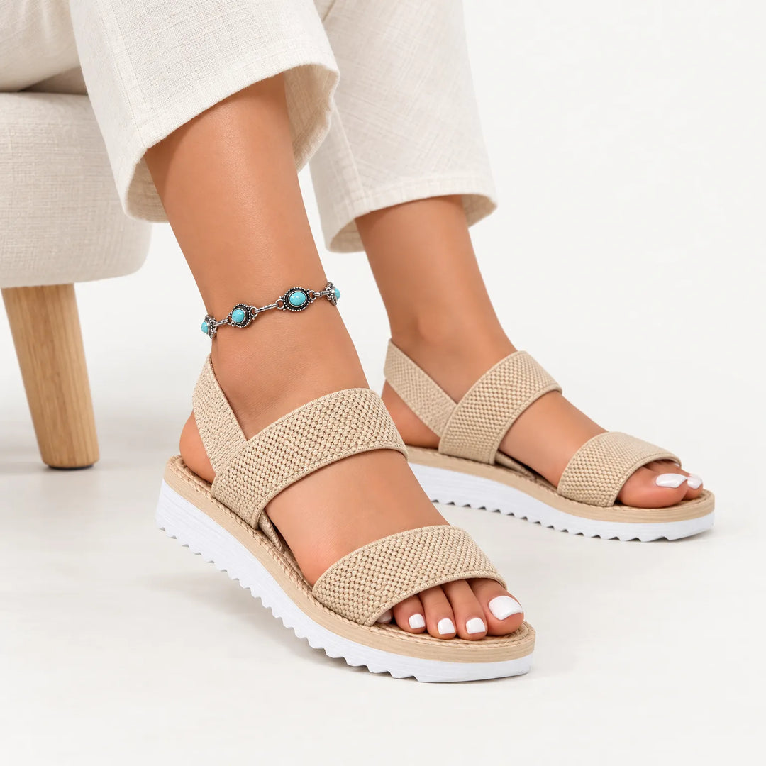 Beige woven elastic double-strap sandals with white ridged soles, worn seated with cream linen pants and a turquoise anklet 1