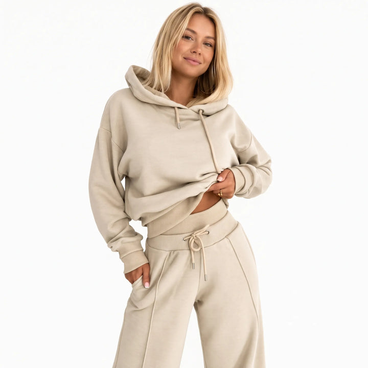 Smiling model in a beige hooded sweatshirt and wide-leg pants against a plain white background 1