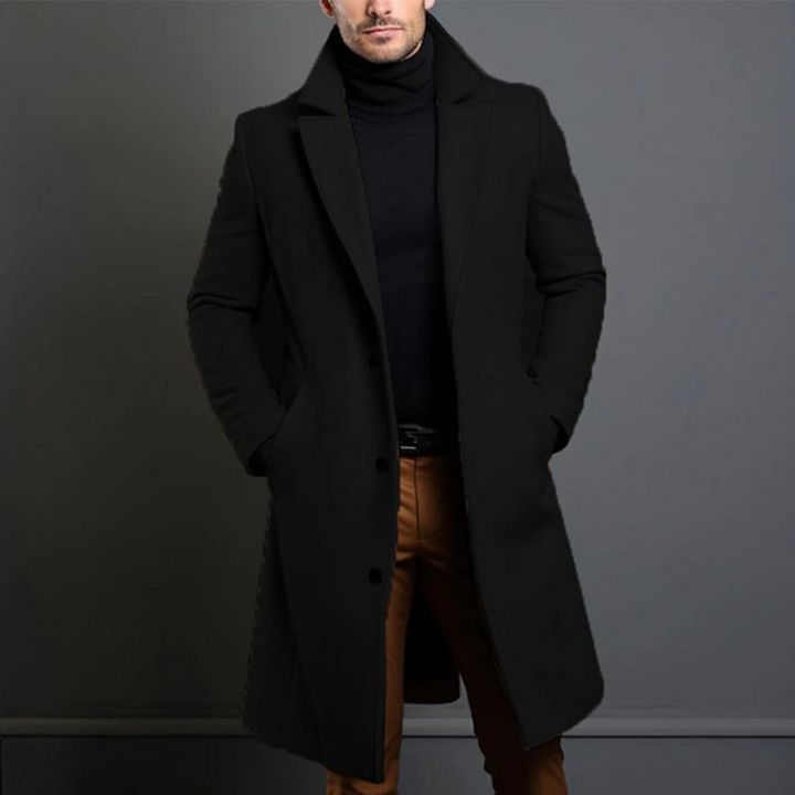 Black single-breasted longline coat with notch lapels, worn open over a black turtleneck and brown trousers, hands in pockets, cropped at the chin. 1