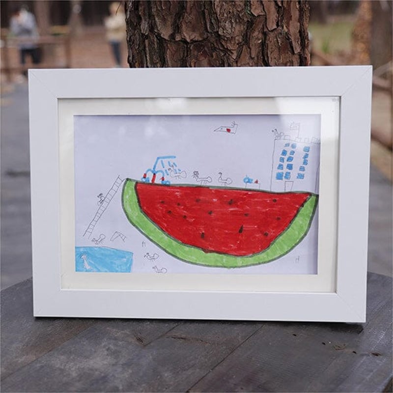 White frame standing on a wooden table outdoors, displaying a child's marker drawing of a giant watermelon slice with tiny people and a building. 21