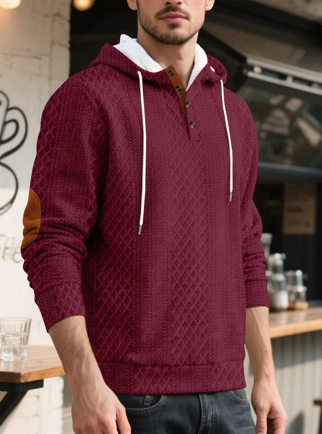 Man wearing a burgundy diamond-textured hoodie with white drawstrings and brown elbow patch, standing near a cafe counter 20