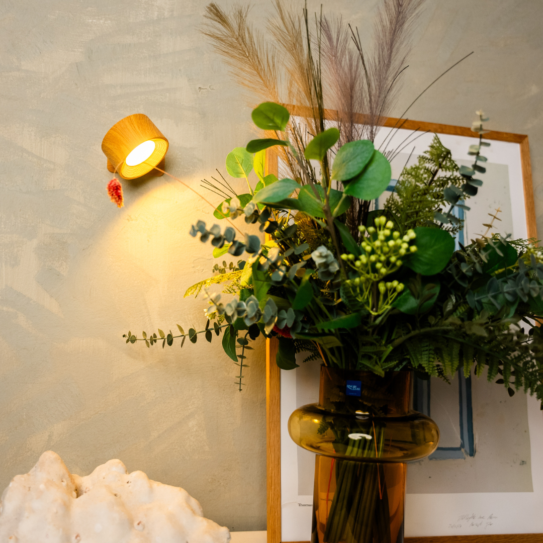 Black wireless LED wall light, another view, beside a framed print and vase arrangement.