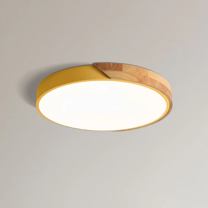 Yellow round ceiling light with a natural wood section on the rim, mounted on a light ceiling and glowing white.