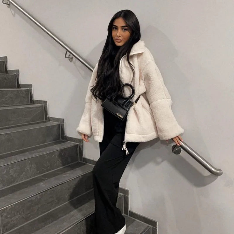 Woman leaning on a stairwell railing in a cream fleece coat, black pants and a small black handbag 2