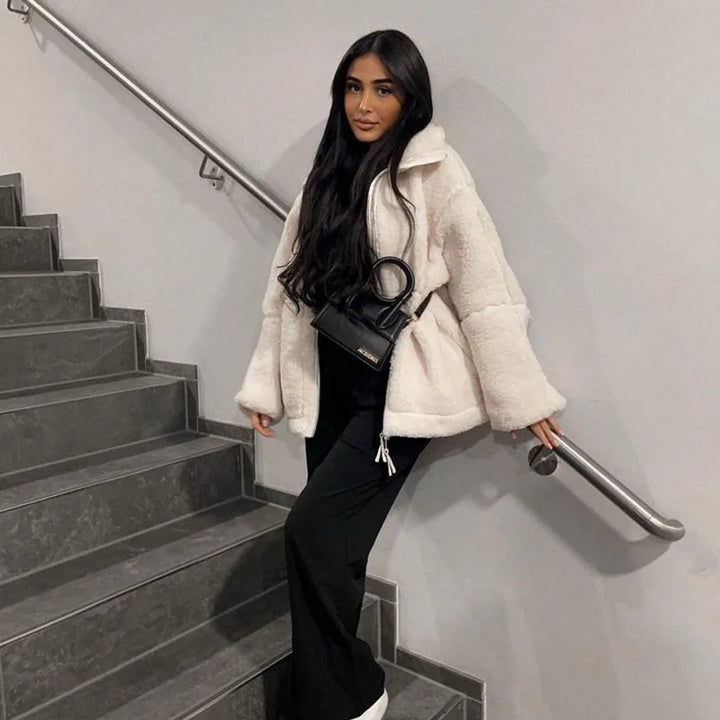 Woman leaning on a stairwell railing in a cream fleece coat, black pants and a small black handbag 2