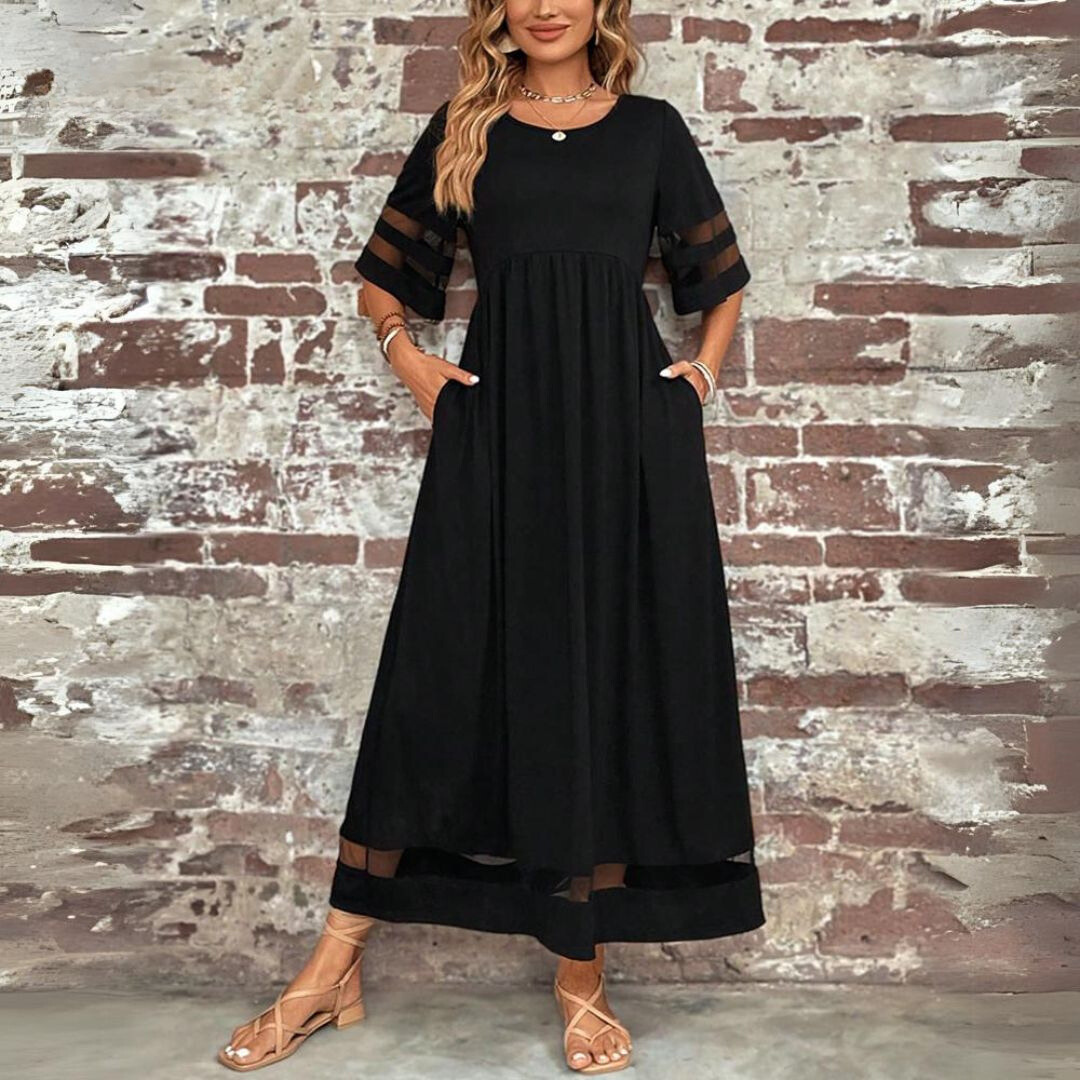 Front view of the black maxi dress with gathered waist, side pockets and sheer-striped sleeves, styled against a brick wall. 2