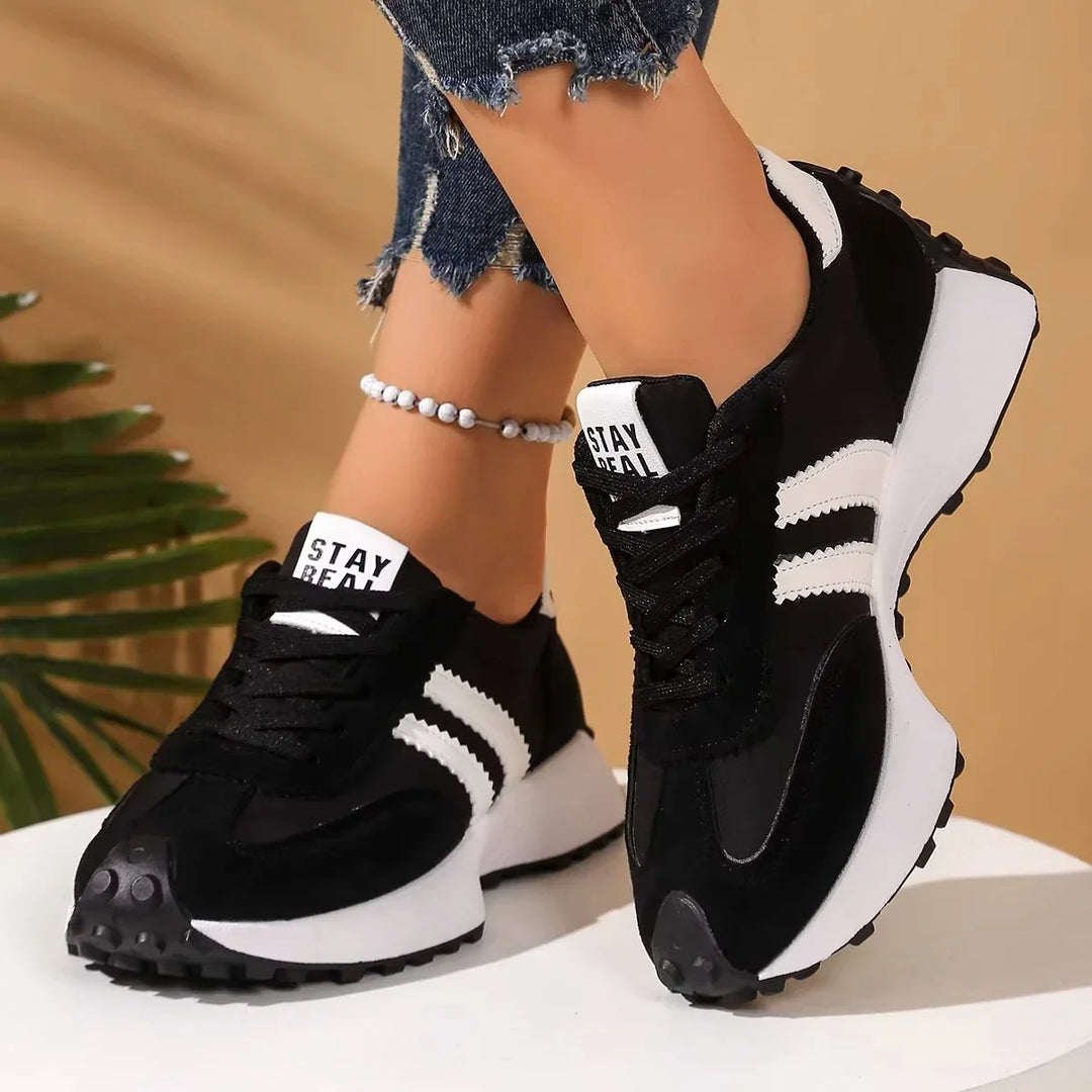 Black platform trainers with white side stripes, worn crossed on a white pedestal beside a palm leaf 2