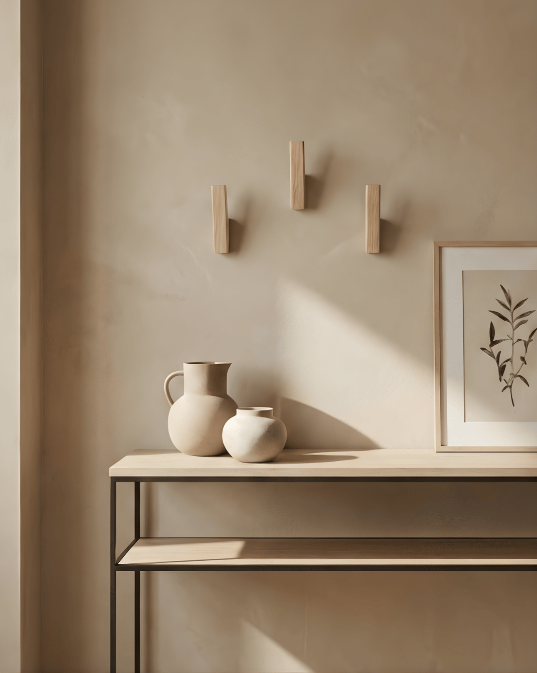 Three light oak wall hooks mounted in a triangle above a slim console table with cream ceramic vases and a framed botanical print. 6