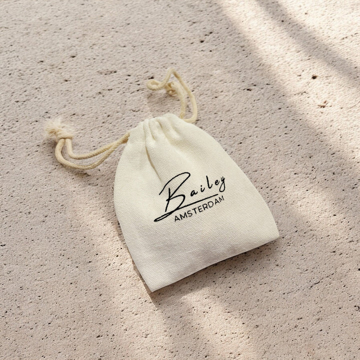 Cream cotton drawstring pouch with black script lettering resting on a sandy, sunlit surface 5