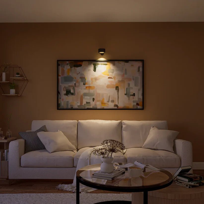 Black wall light above a framed abstract painting, lighting the art over a cream sofa.